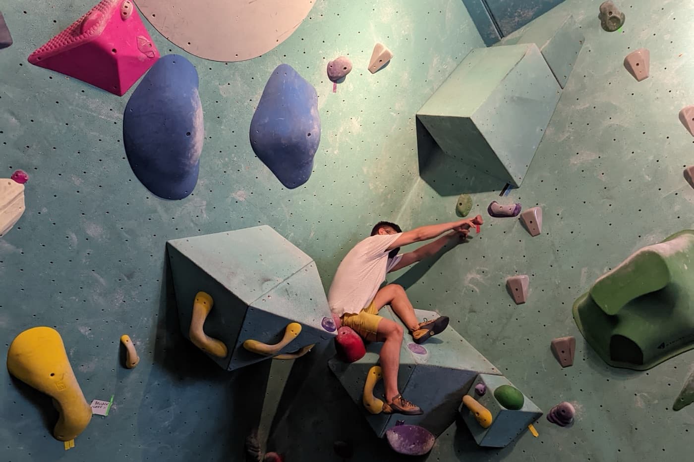 A climber in a white shirt on a steep bouldering gym wall, reaching for a small hold between large coloured volumes.