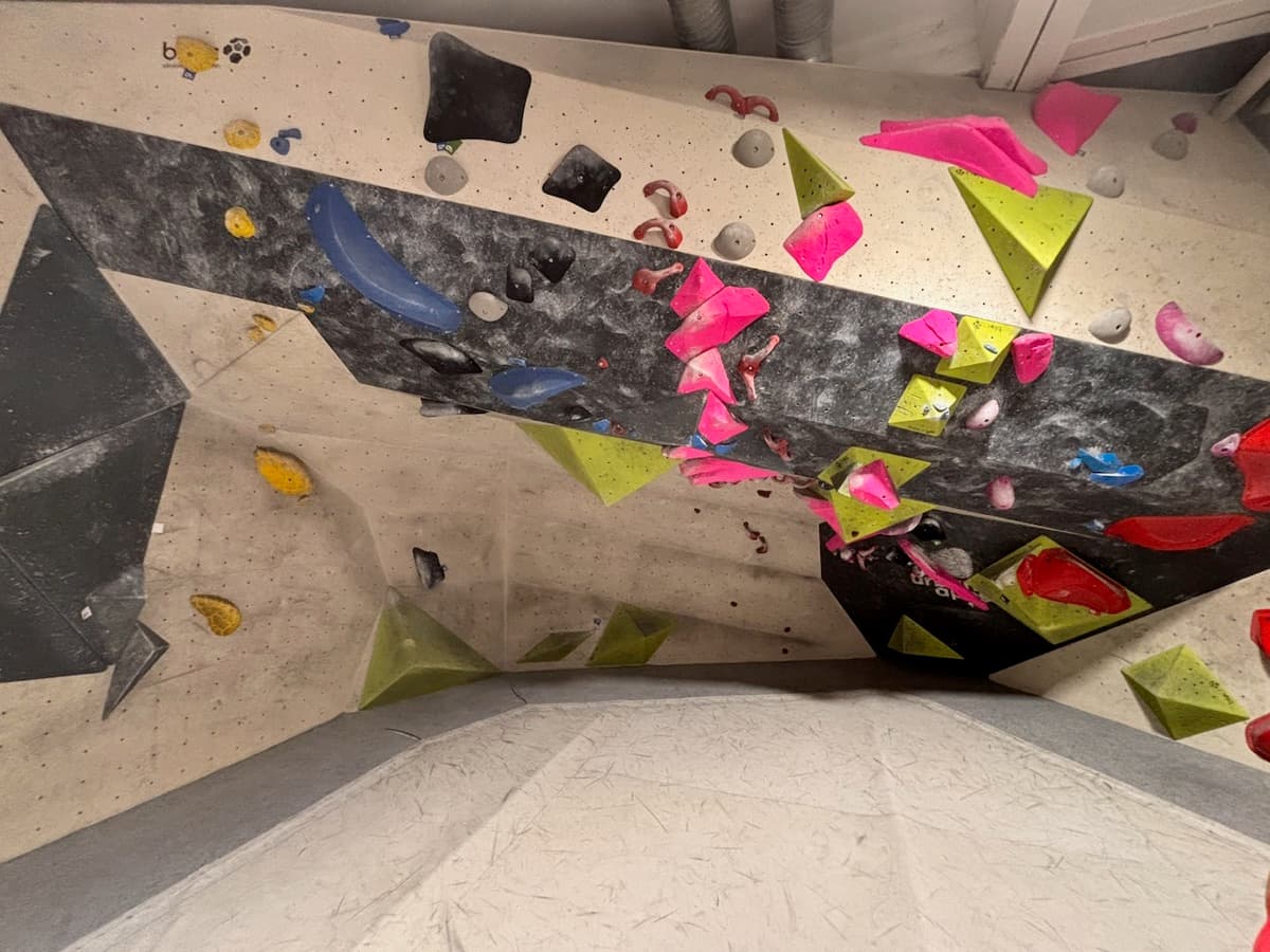An overhanging bouldering wall in the gym, seen from below, with pink, green, blue and black holds and several large volumes.