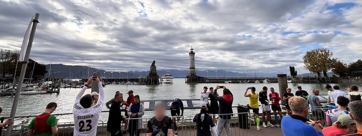 Lindau harbour on Lake Constance under a wide cloudy sky with patches of blue: the stone lion and the white lighthouse at the harbour entrance, a passenger boat between them, mountains on the far shore, and in the foreground runners in race kit at the railing, several of them taking photos of the harbour.