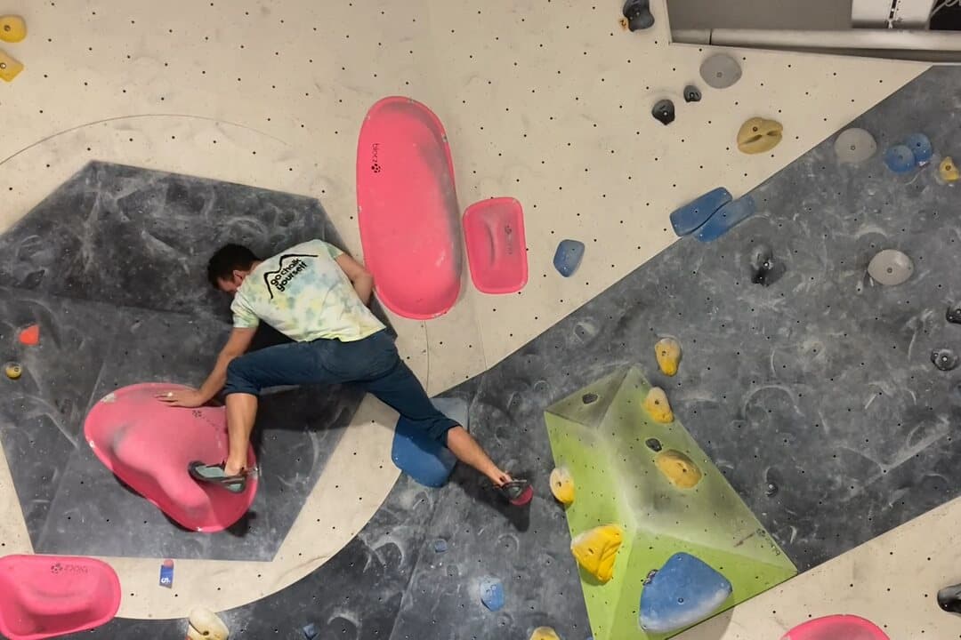 A boulderer in a t-shirt and shorts moving across a colourful indoor bouldering wall.