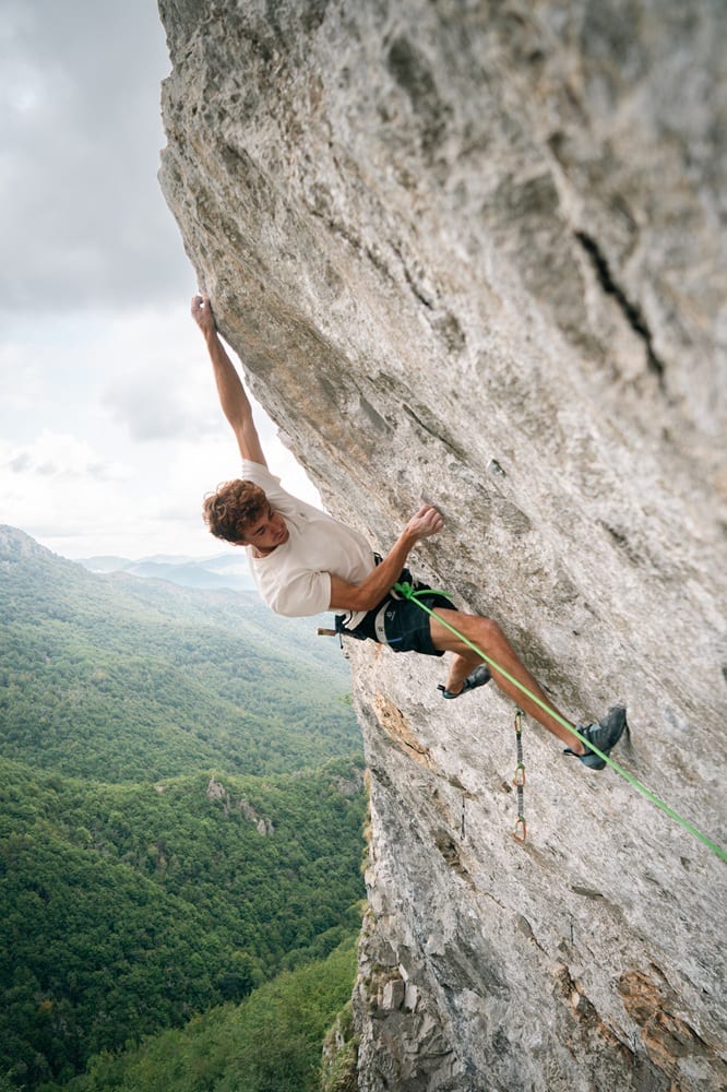 Filip Schenk reaching up on a steep grey limestone wall above a wooded valley