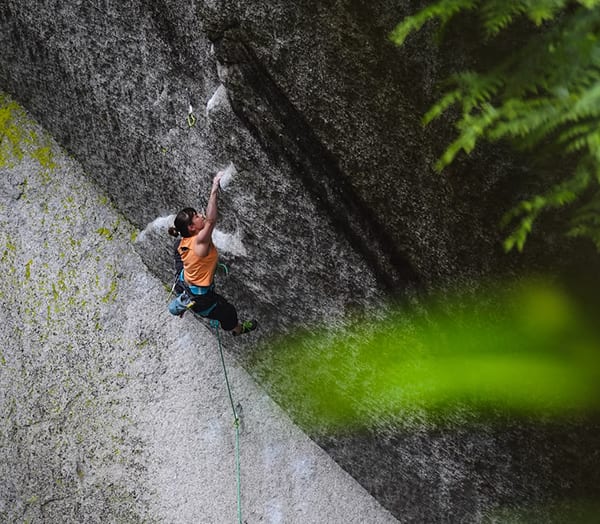Climber in an orange top on a steep granite face in Squamish with ferns in the foreground