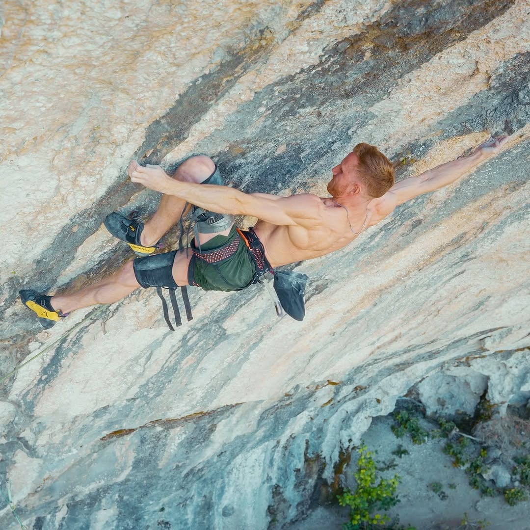 Shirtless climber spanning wide across a streaked grey and blue limestone wall