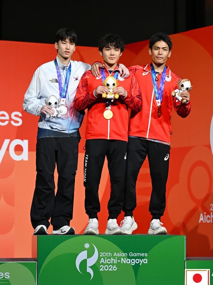Three men holding medals on the Asian Games podium in Nagoya, the winner in the centre in a red Team Japan jacket