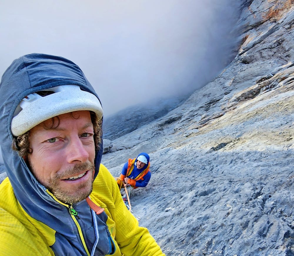 Selfie of a climber in a helmet with his partner on the grey north face below