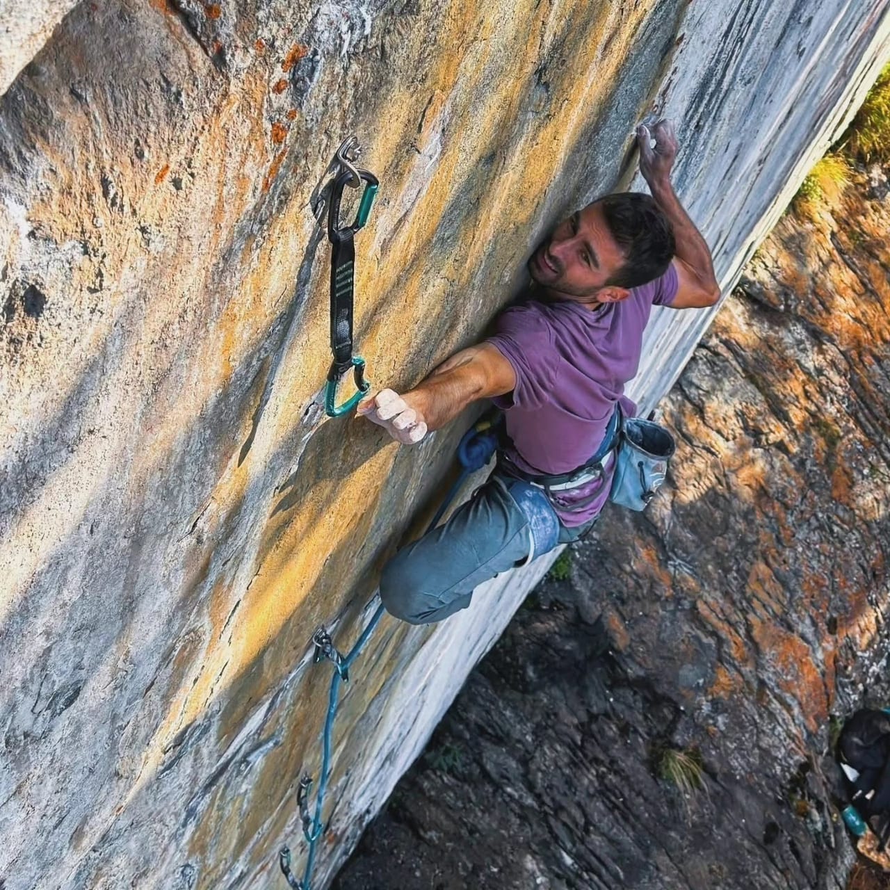 A climber in a purple shirt reaching past a quickdraw on orange streaked limestone